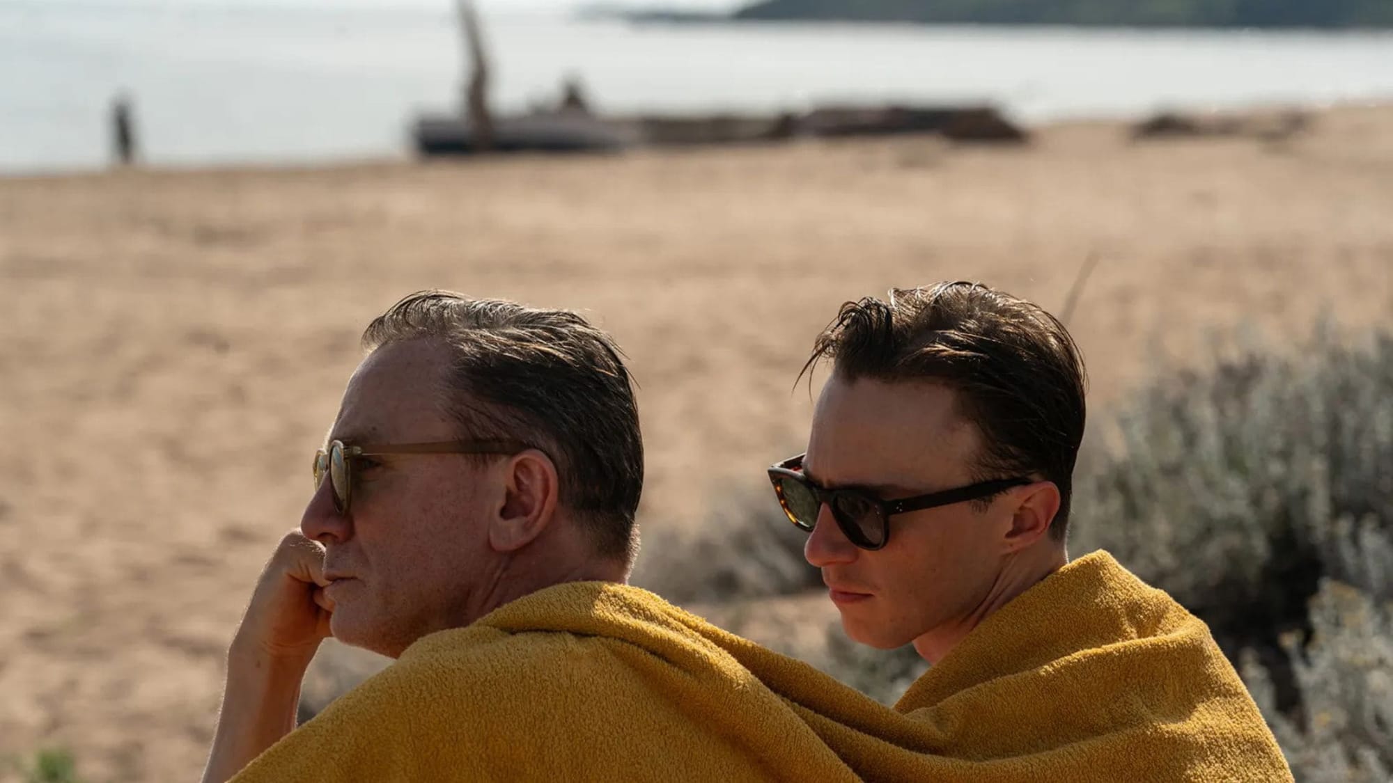 Primer plano de perfil de dos hombres con gafas de sol sentados en una playa de arena, compartiendo una gran toalla amarilla sobre sus hombros. Al fondo se aprecia el mar difuminado.