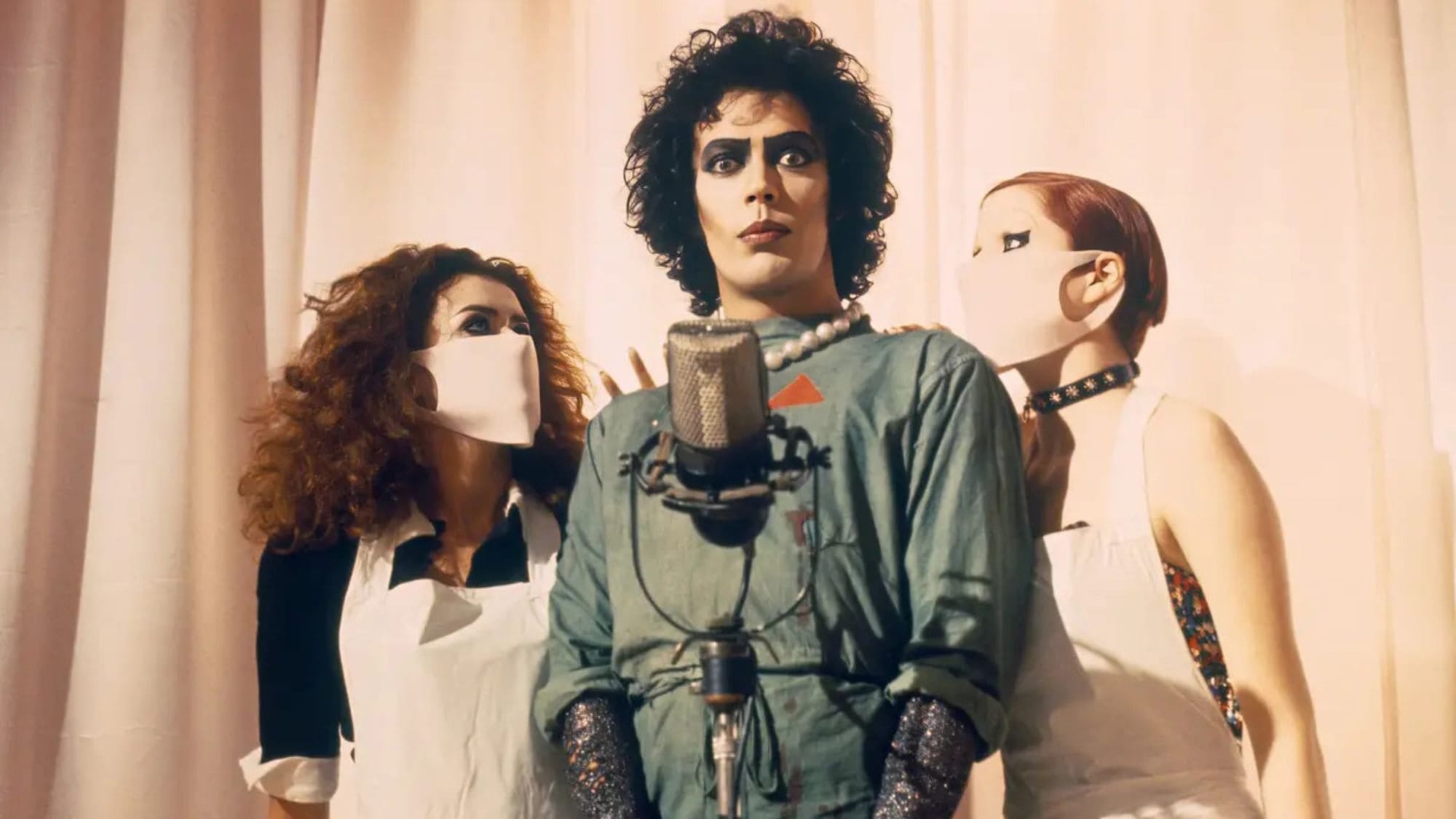 Tim Curry como el Dr. Frank-N-Furter con maquillaje dramático de ojos y labios frente a un micrófono clásico de plata, flanqueado a los lados por Columbia y Magenta usando mascarillas quirúrgicas blancas sobre un fondo claro.